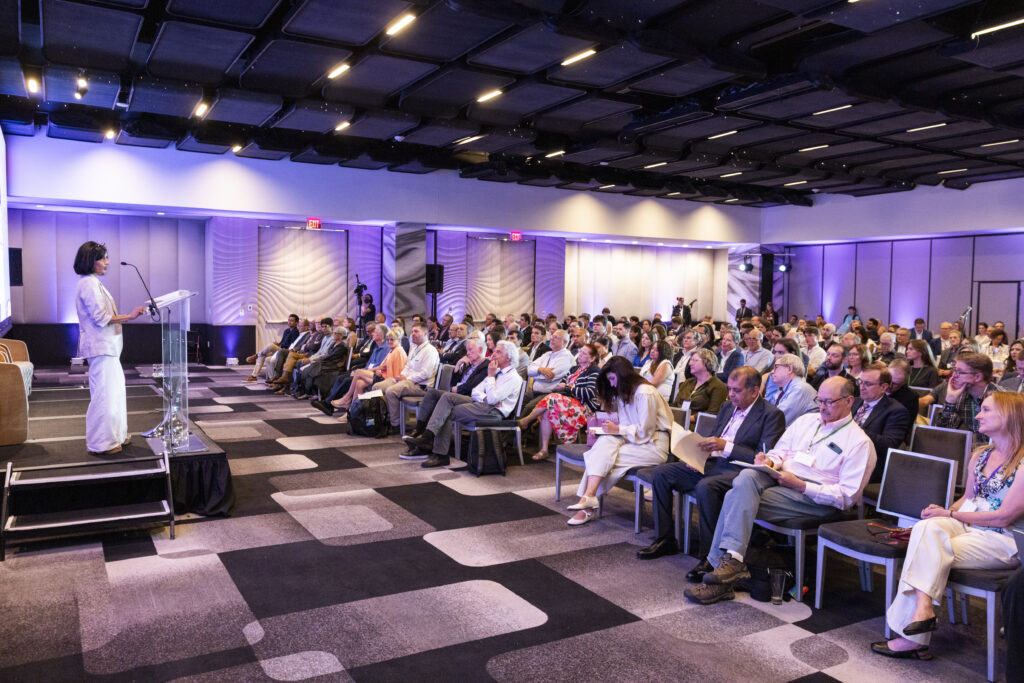 A woman speaks to a hotel ballroom full of people.